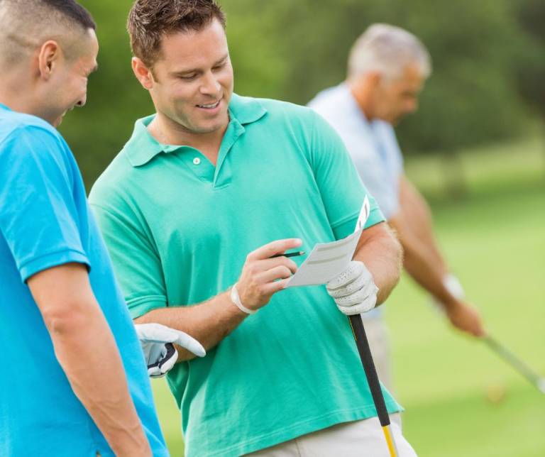 A golfer in a teal polo reviews his scorecard with another golfer in a baby blue polo.